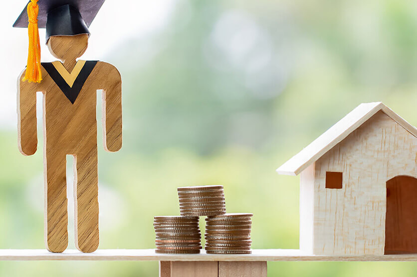 Wooden figure with black graduation hat balancing next to stacks of quarters and wooden house.